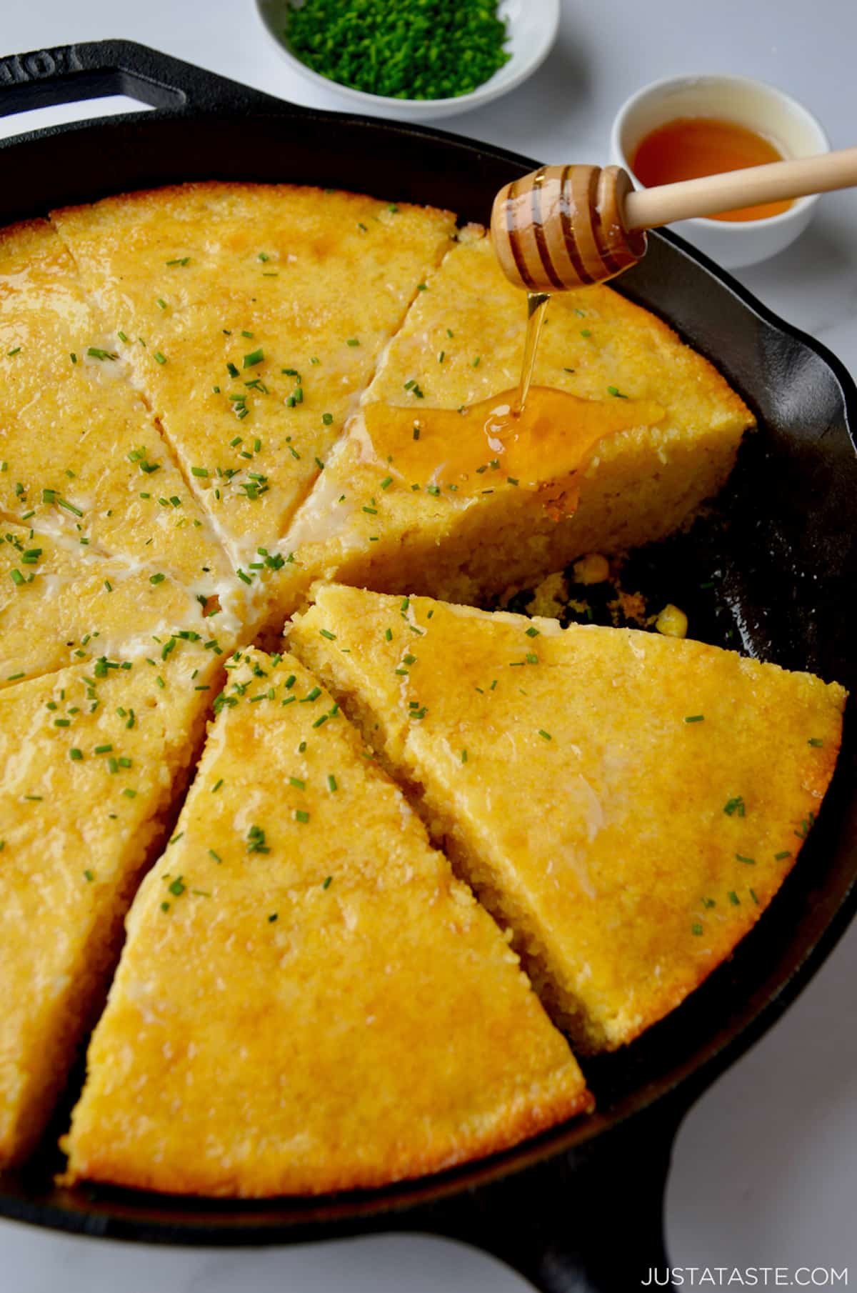Honey being drizzled over sliced cornbread in a cast iron pan. The cornbread is topped with butter and minced chives and small bowls of more chives and honey are beside the skillet.
