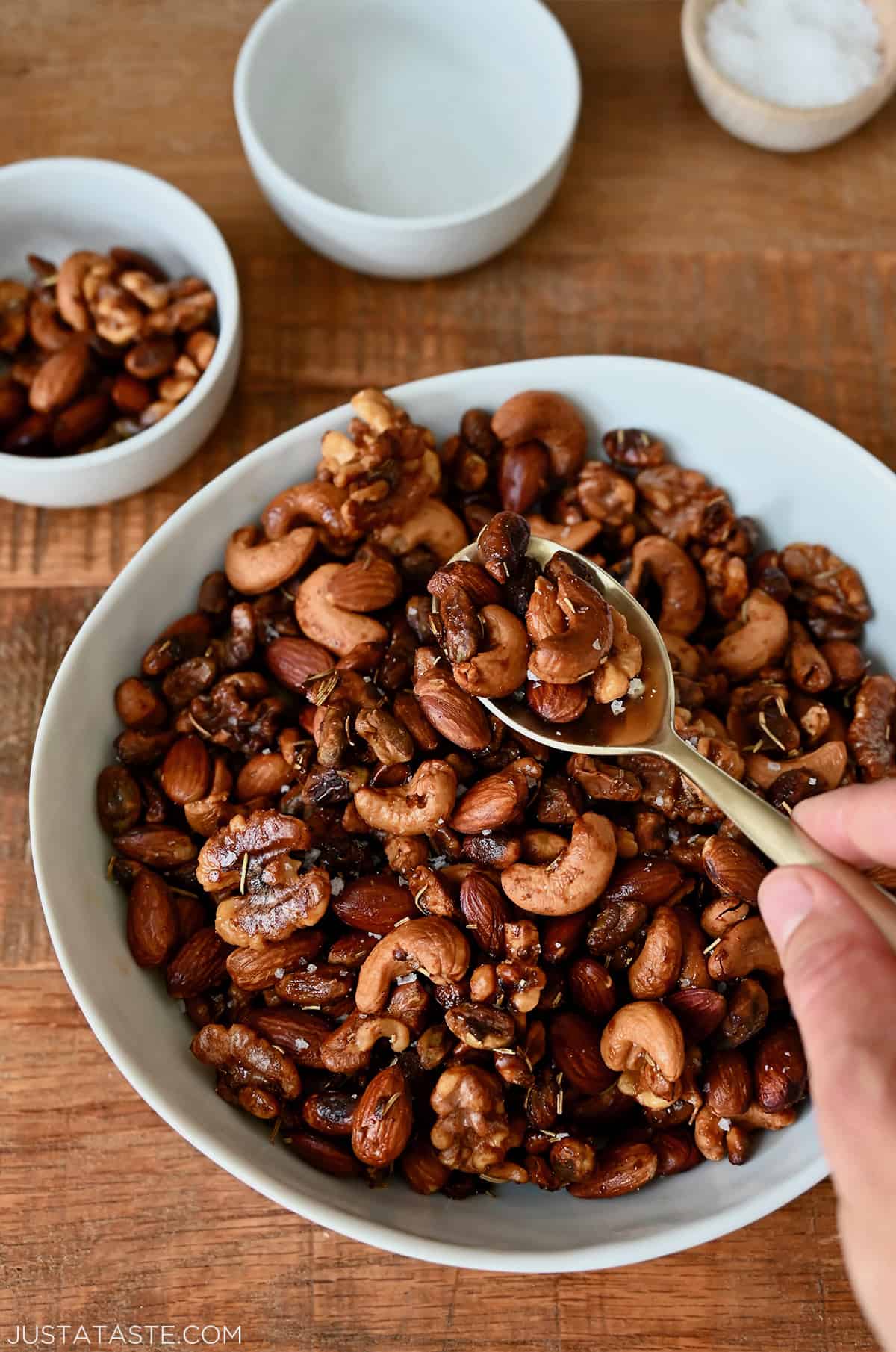 A person scooping roasted nuts out of a white bowl. Two other bowls and a small bowl of salt are behind the nuts.