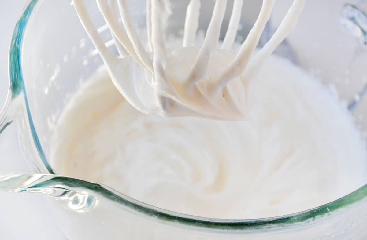 Whipped cream in the bowl of a stand mixer fitted with the whisk attachment.