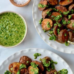 Two plates of roasted potatoes topped with chimichurri sauce. Small bowls of chimichurri sauce and flakey salt sit beside the plates.