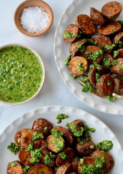 Two plates of roasted potatoes topped with chimichurri sauce. Small bowls of chimichurri sauce and flakey salt sit beside the plates.