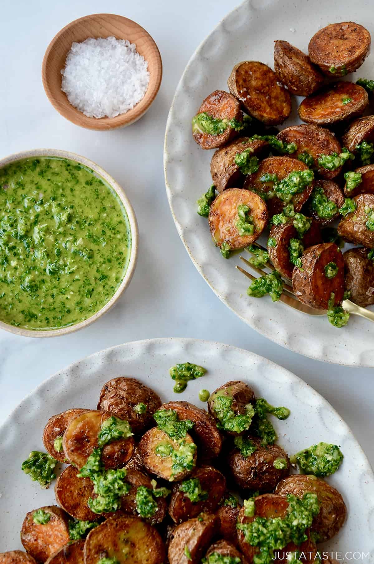 Two plates of roasted potatoes topped with chimichurri sauce. Small bowls of chimichurri sauce and flakey salt sit beside the plates.