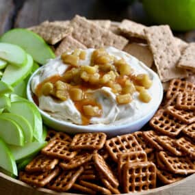 A bowl of cheesecake dip topped with caramel apples surrounded by pretzels, sliced apples and graham crackers. Two green apples are in the background.