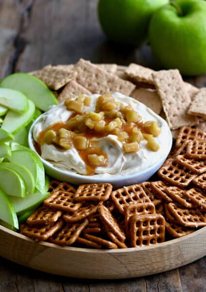 A bowl of cheesecake dip topped with caramel apples surrounded by pretzels, sliced apples and graham crackers. Two green apples are in the background.