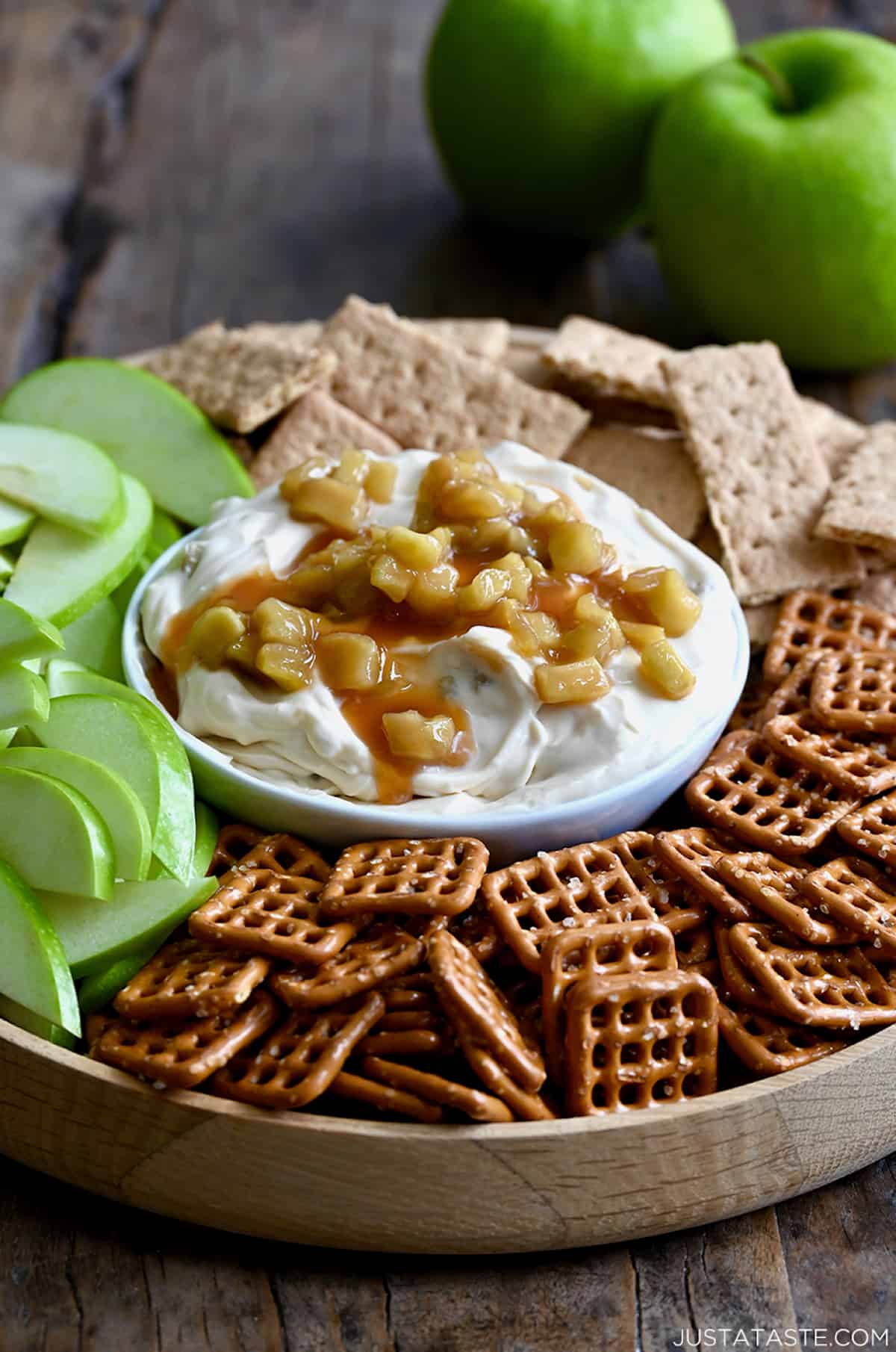 A bowl of cheesecake dip topped with caramel apples surrounded by pretzels, sliced apples and graham crackers. Two green apples are in the background.