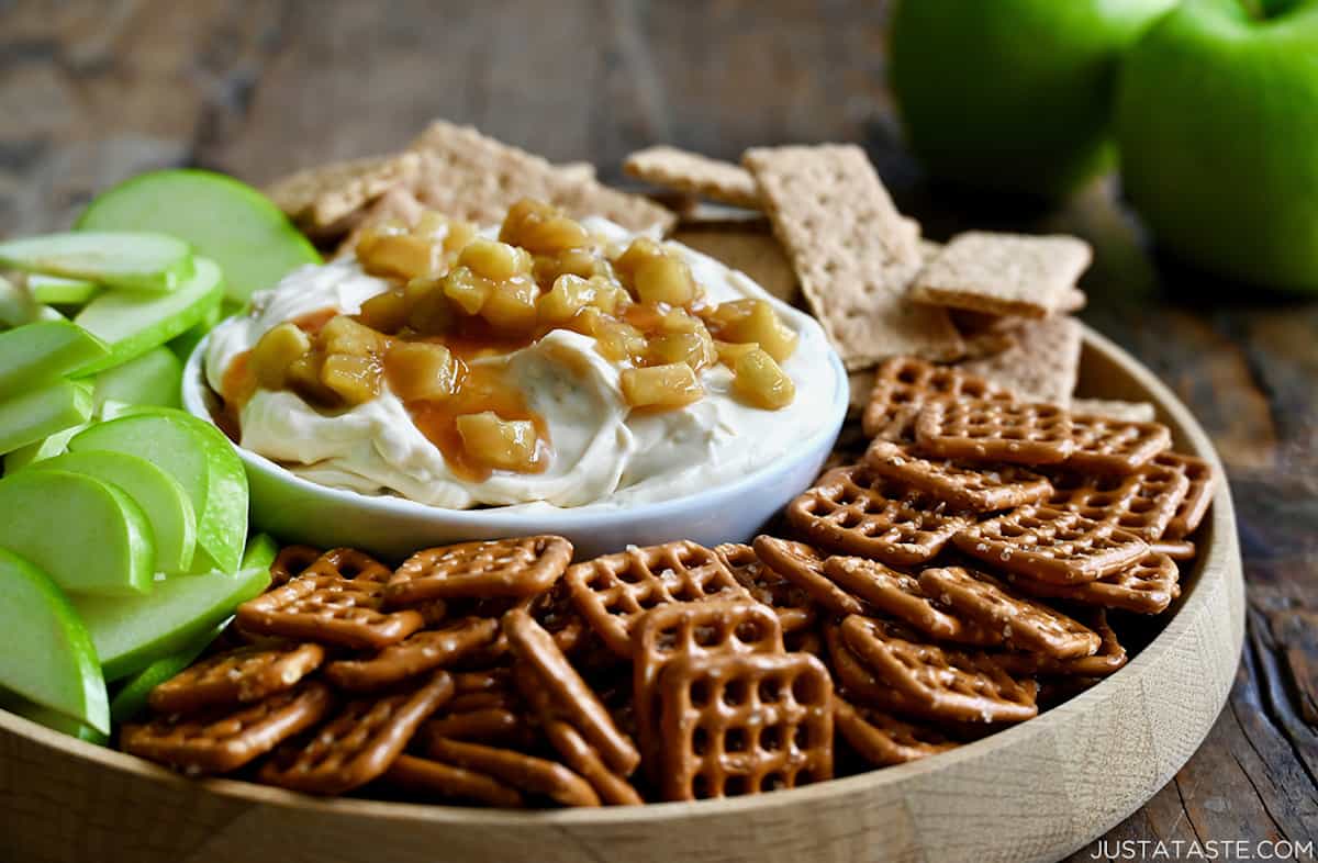 A platter with a bowl of cheesecake dip topped with sautéed apples in its center. Pretzels, sliced apples and graham crackers surround the dip. Two apples sit behind the platter.