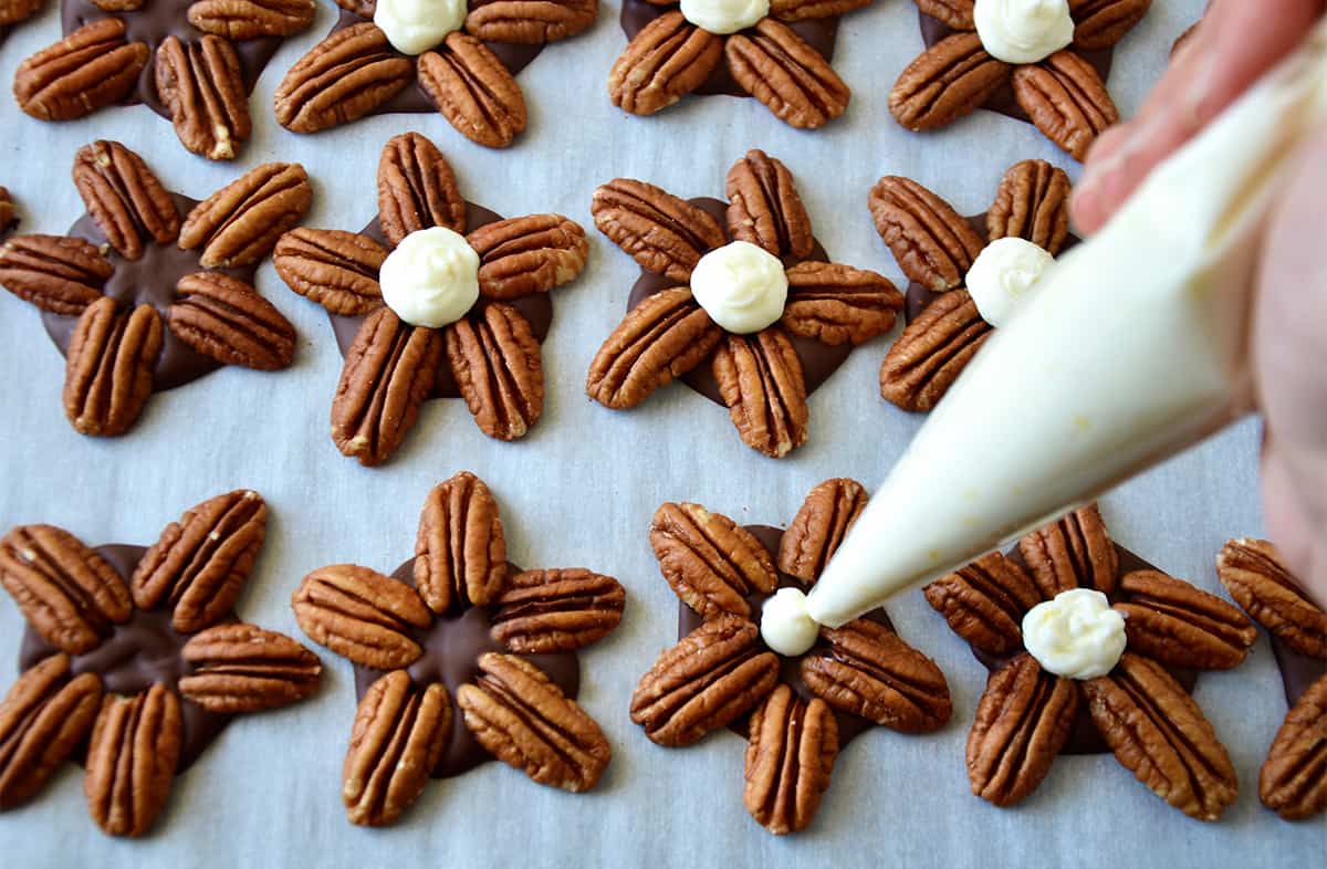 A person using a piping bag to add cheesecake filling to chocolate turtle candies on a parchment-lined baking sheet.