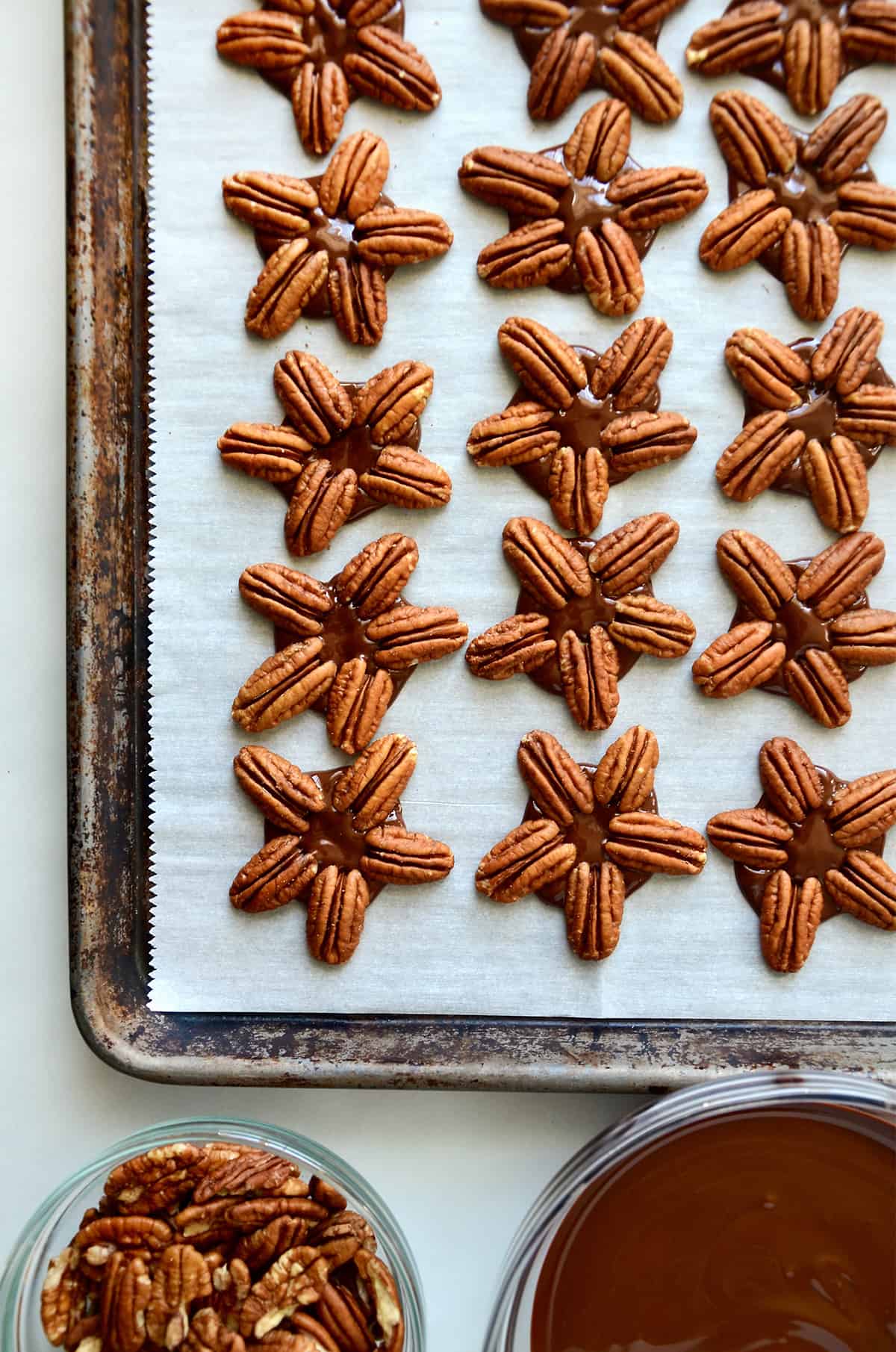 Chocolate turtles on a parchment-lined baking sheet with bowls of pecan halves and melted chocolate beside the baking sheet.