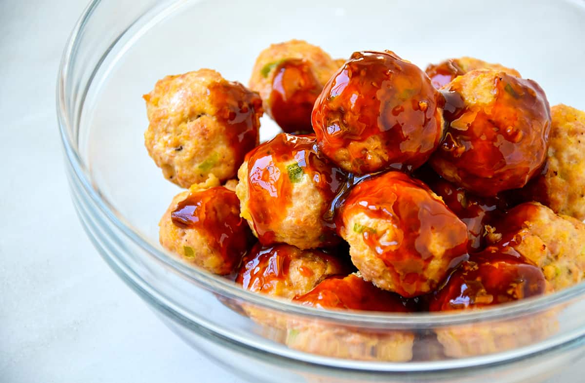 Baked chicken meatballs in a large glass mixing bowl with teriyaki sauce poured on top of them.