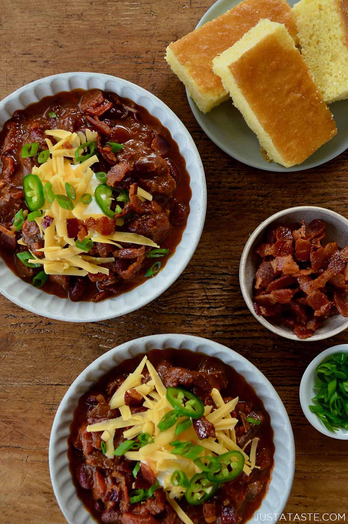 Two bowls of chili topped with sour cream, crumbled bits of bacon, grated cheese, jalapeno slices and thinly sliced scallions. A plate of cornbread and small bowls containing more bacon and scallions sit beside the bowls of chili.