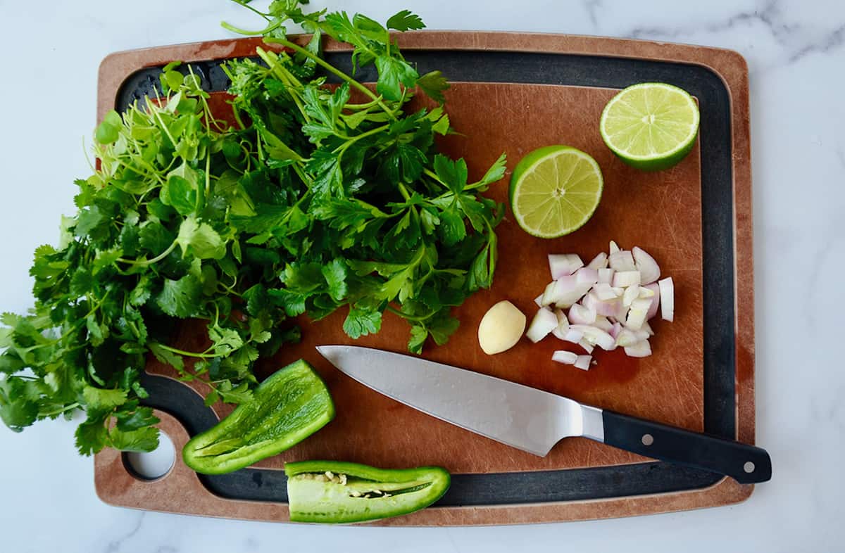 Cilantro, parsley, a halved jalapeño pepper, a peeled clove of garlic, chopped shallots and a halved lime on a cutting board.