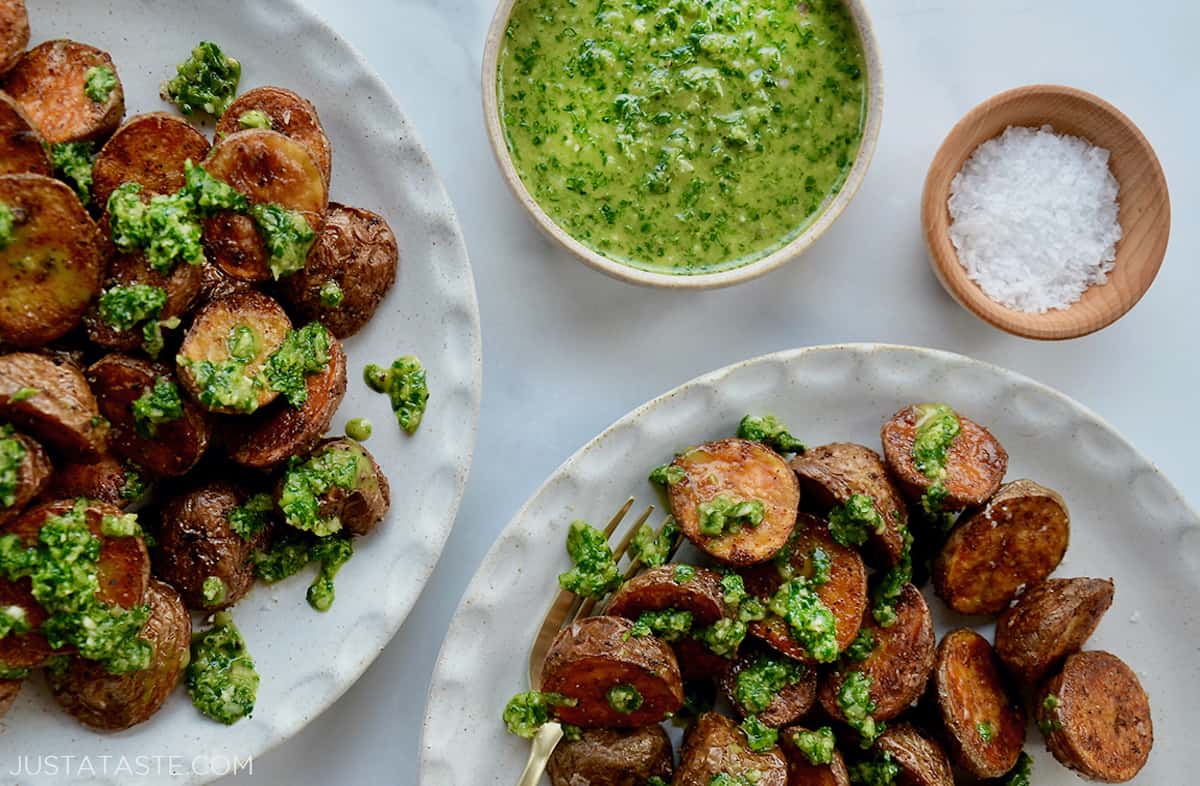 Two plates of roasted potatoes drizzled with chimichurri sauce. Small bowls of chimichurri sauce and flakey salt sit next to the plates.