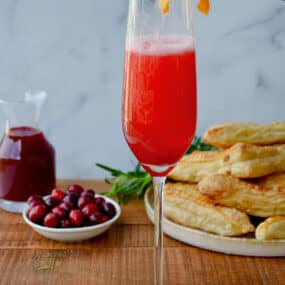 A person pouring Prosecco into a champagne flute with cranberry syrup. A plate with puff pastry turnovers, a bowl of cranberries and a carafe of cranberry syrup are behind the flute.