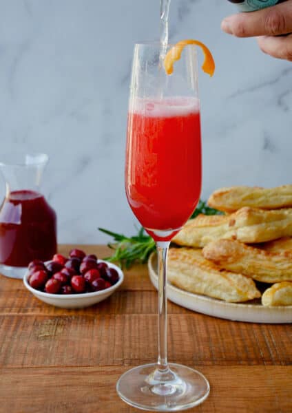 A person pouring Prosecco into a champagne flute with cranberry syrup. A plate with puff pastry turnovers, a bowl of cranberries and a carafe of cranberry syrup are behind the flute.