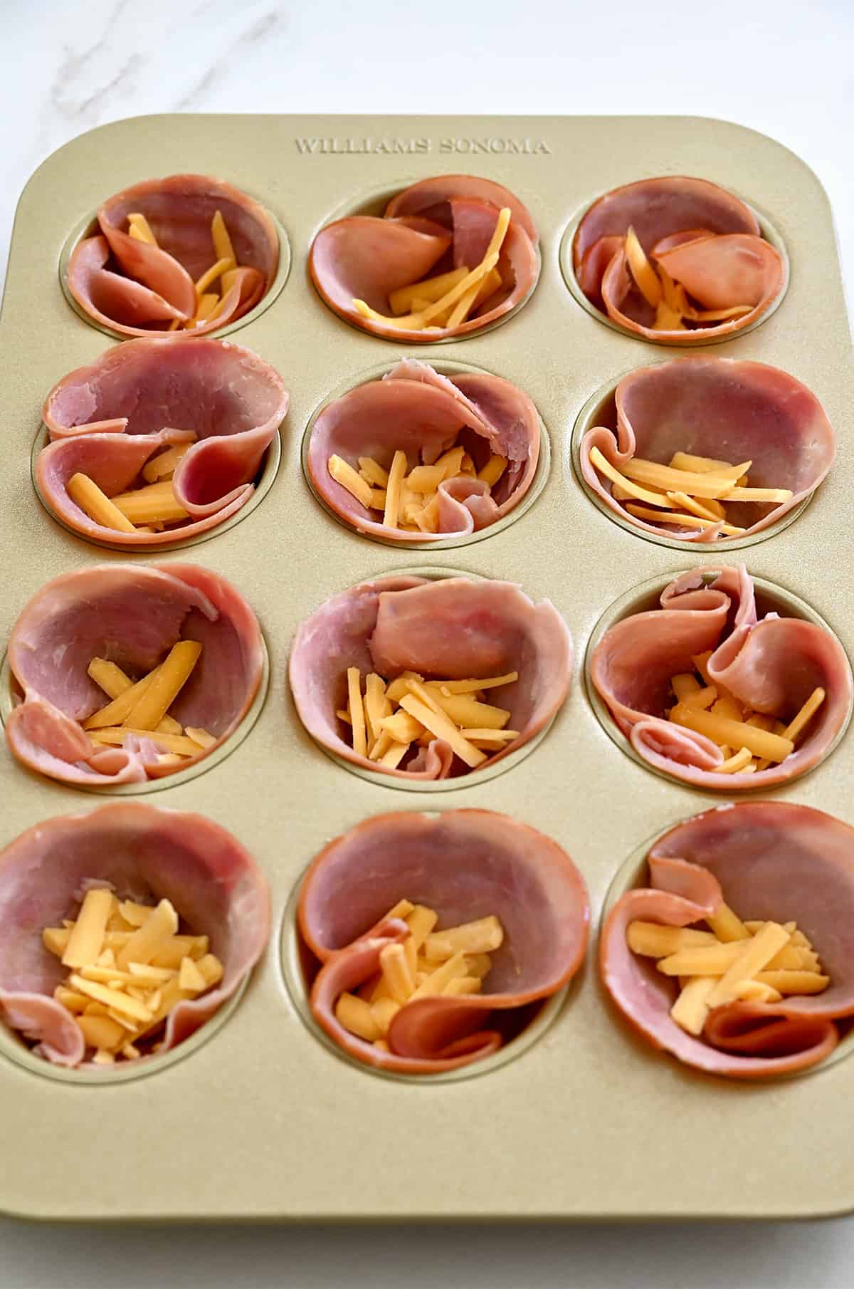 A muffin tin containing thin slices of ham topped with shredded cheddar cheese.