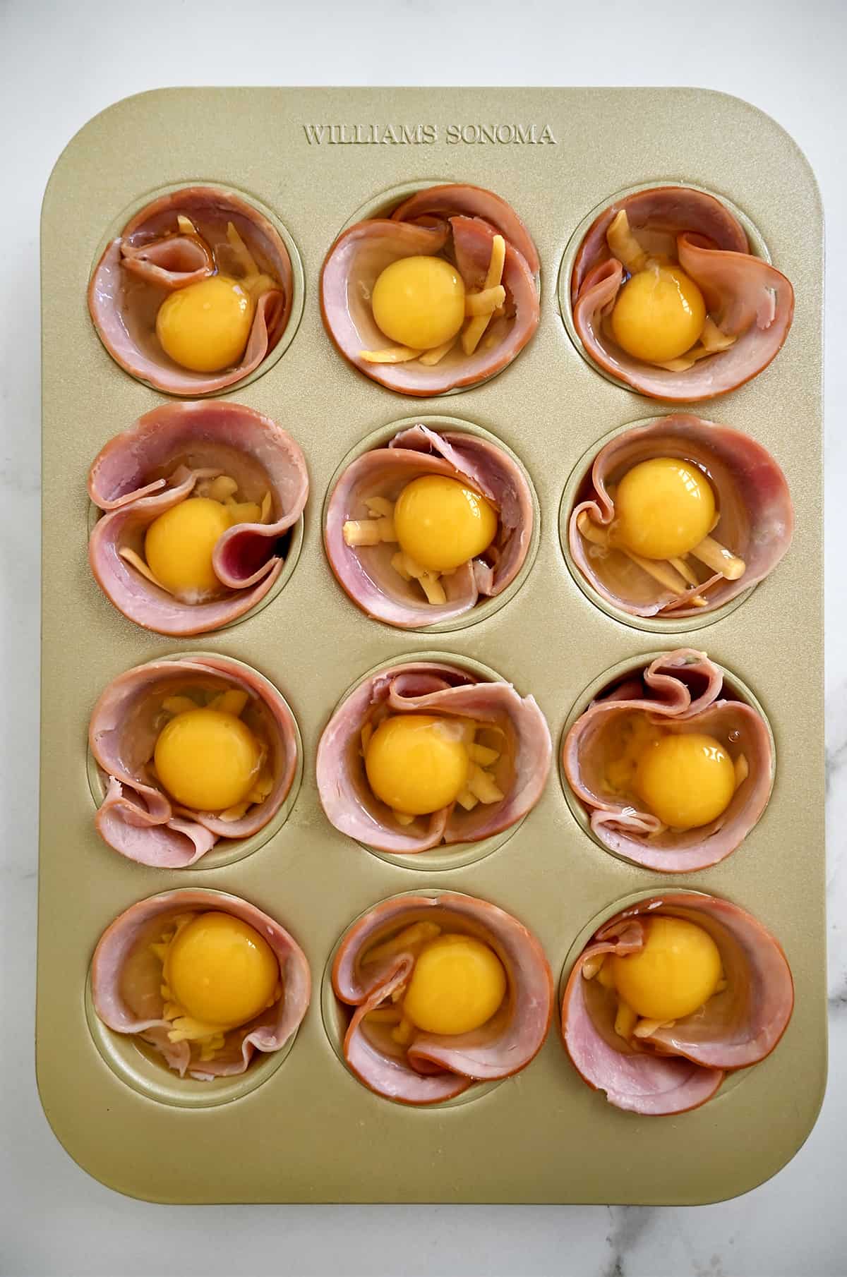 A muffin pan containing a thin slice of ham in each cup along with an uncooked egg.