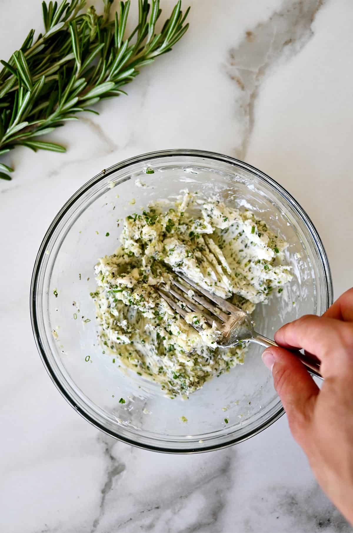 A hand holding a fork mixes together softened butter with garlic and herbs in a bowl.