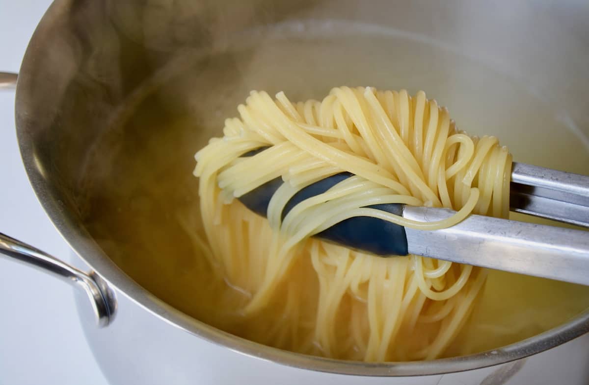 A pair of tongs being used to lift cooked spaghetti out of boiling water.