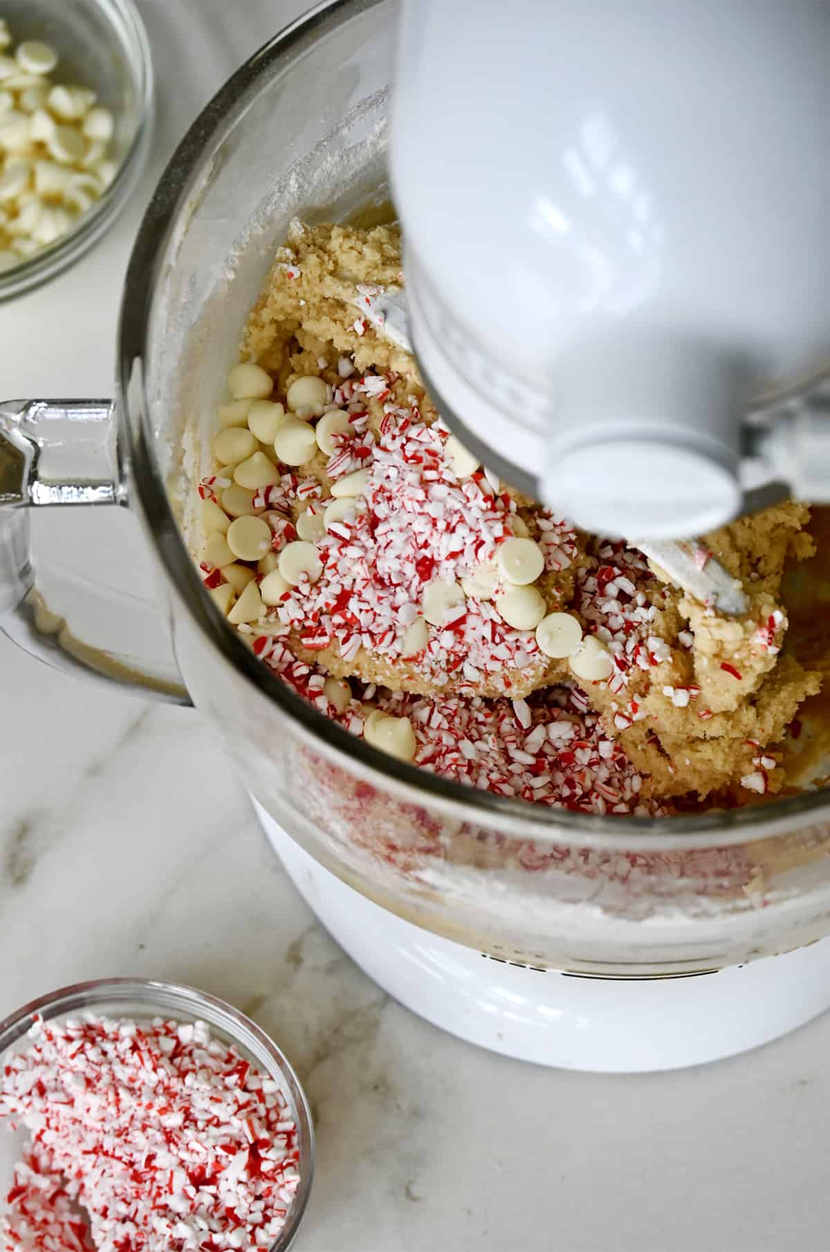A white stand mixer with a clear bowl containing cookie dough topped with crushed candy canes and white chocolate chips. A small bowl filled with crushed candy canes is next to it.