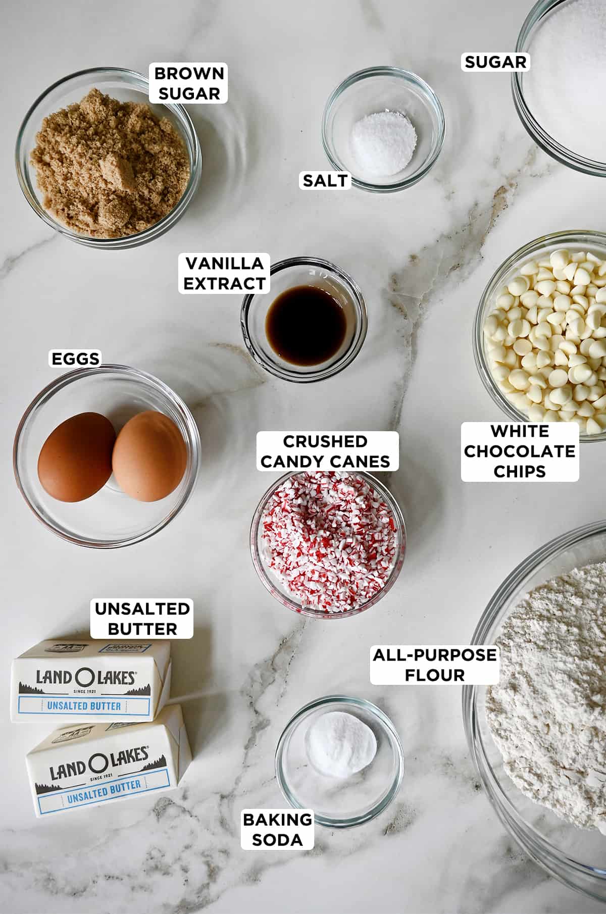 Various sizes of clear bowls containing the ingredients needed to make peppermint white chocolate cookies, including light brown sugar, salt, white sugar, vanilla extract, white chocolate chips, crushed candy canes, all-purpose flour, baking soda, butter and eggs.