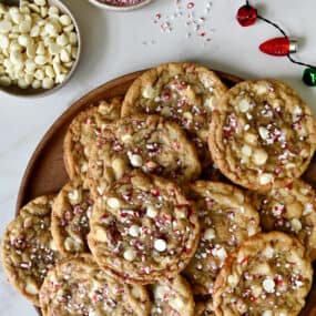 White chocolate peppermint cookies on a platter next to a two small bowls containing white chocolate chips and crushed candy canes.