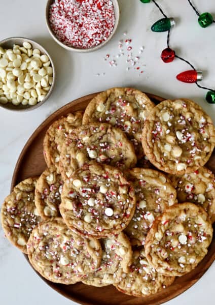 White chocolate peppermint cookies on a platter next to a two small bowls containing white chocolate chips and crushed candy canes.
