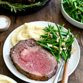 Medium-rare prime rib on a white dinner plate with green beans and mashed potatoes. A fork and knife are also on the plate.