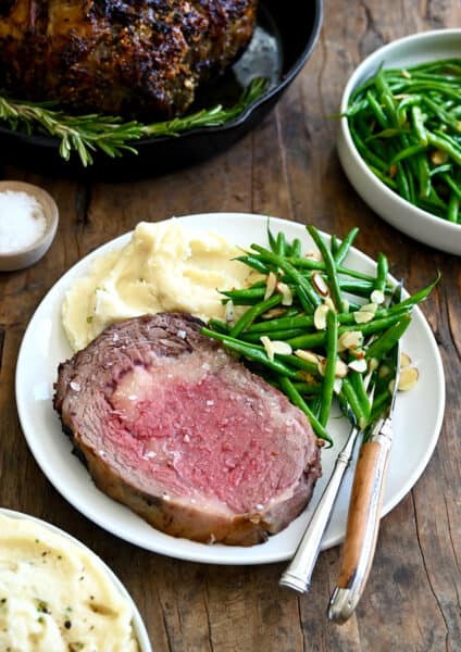 Medium-rare prime rib on a white dinner plate with green beans and mashed potatoes. A fork and knife are also on the plate.