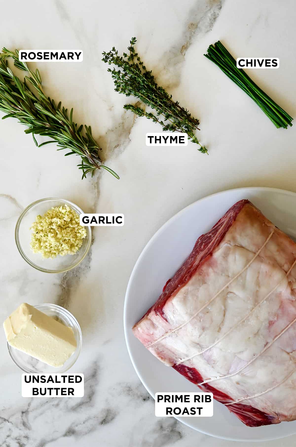 Sprigs of fresh rosemary and thyme next to fresh chives, a prime rib roast on a white plate, unsalted butter in a clear bowl and minced garlic in a bowl. 