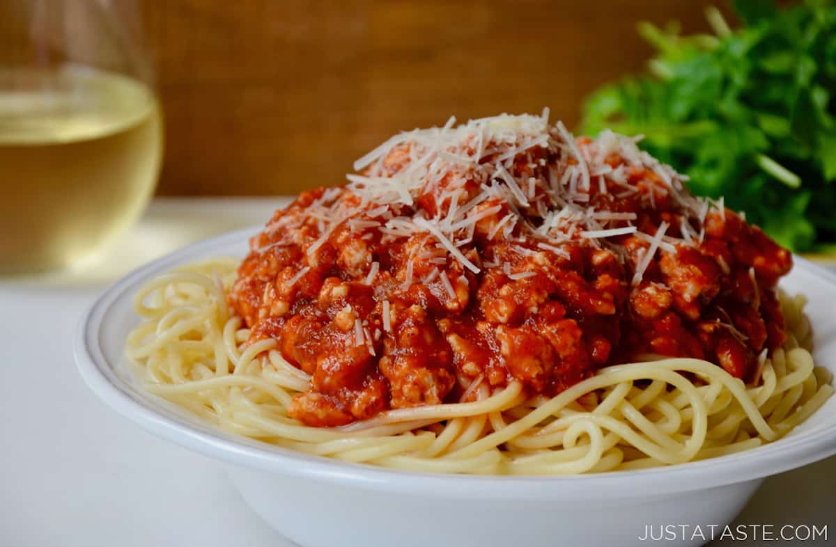 A bowl of spaghetti topped with turkey bolognese and grated Parmesan cheese. A glass of white wine and a salad sit behind the bowl.