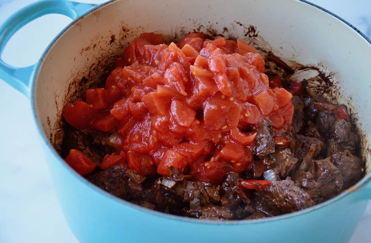 Canned, chopped tomatoes atop browned pieces of beef mixed with vegetables and spices in a Dutch oven.
