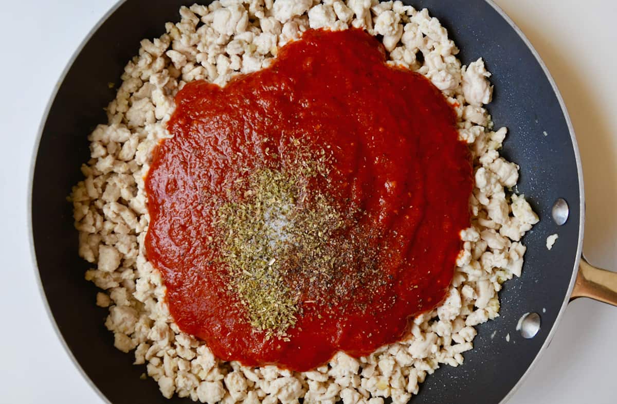 Cooked ground turkey in a nonstick skillet with tomato sauce and spices sitting atop it in the center, ready to be mixed in.