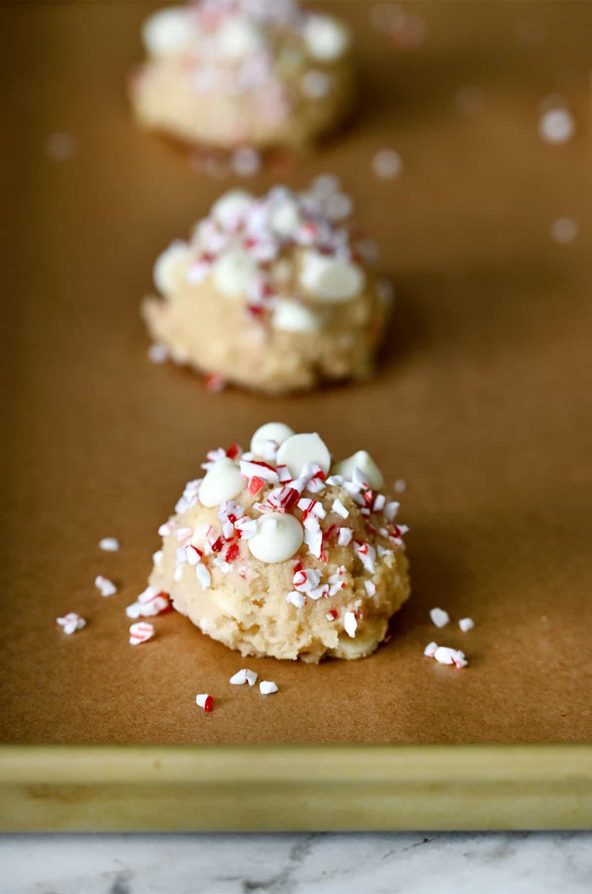 Mounds of peppermint cookie dough topped with white chocolate chips and crushed candy canes on a parchment paper-lined baking sheet.