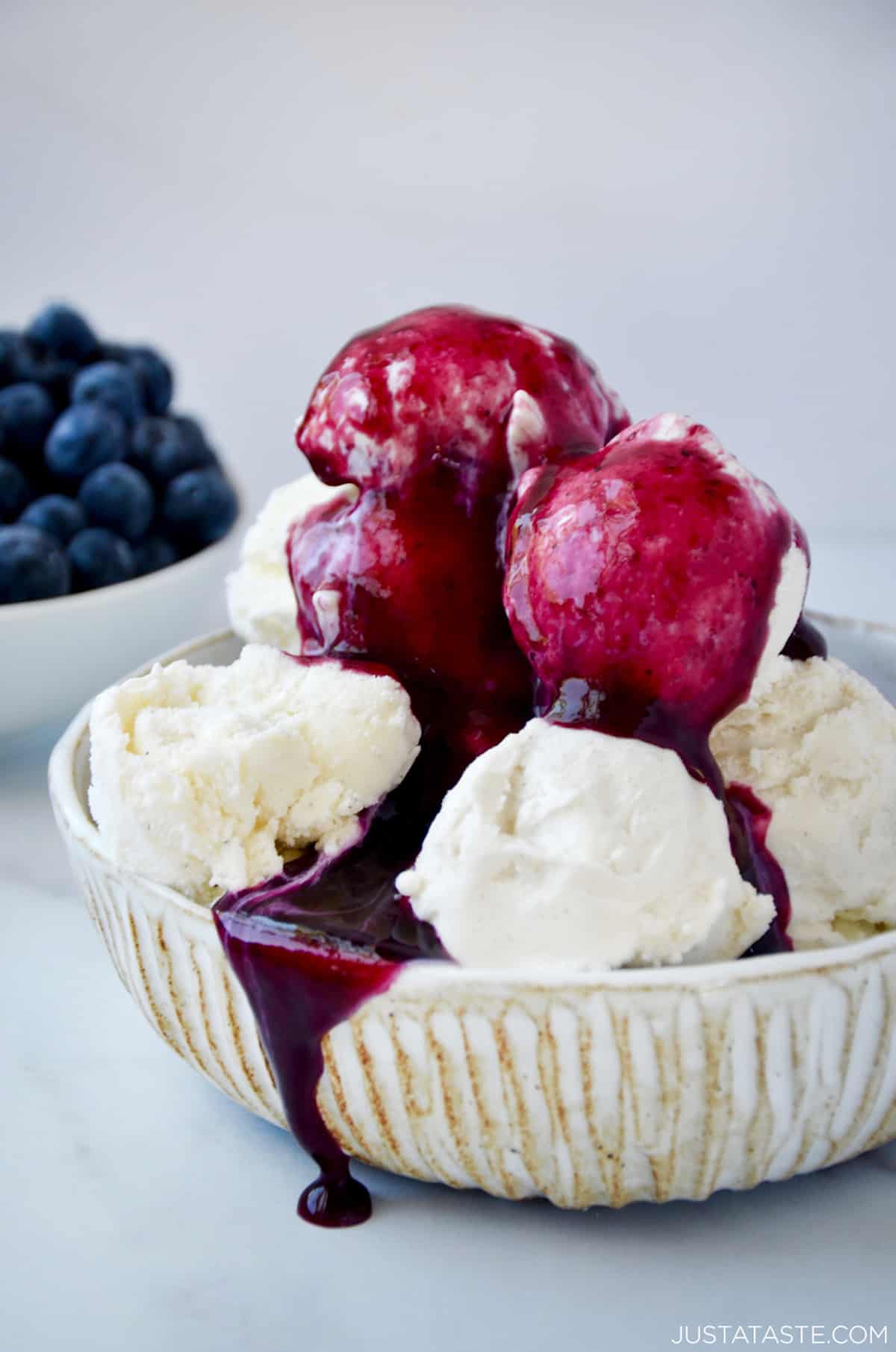 Blueberry sauce poured on top of a bowl of vanilla ice cream. A small bowl containing fresh blueberries sits behind the ice cream.