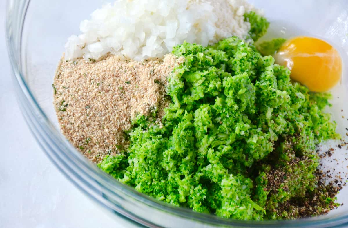 Finely chopped broccoli, salt, pepper, breadcrumbs, onion, grated cheese and an egg in a large glass mixing bowl.