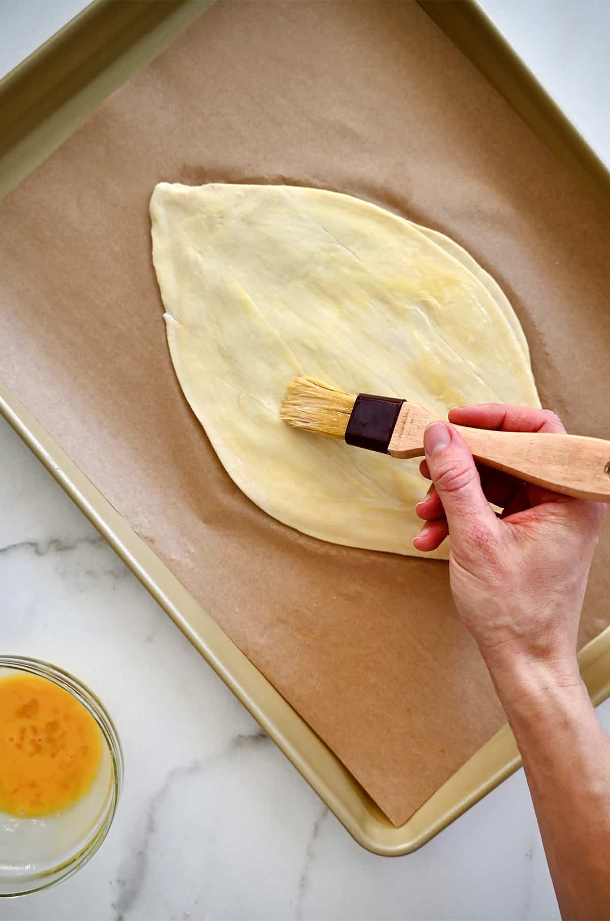 A hand holding a pastry brush applies an egg wash to football-shaped puff pastry on a parchment paper-lined baking sheet.