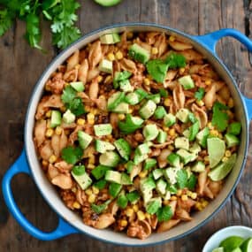 A dutch oven containing cheesy turkey taco pasta topped with diced avocado and chopped fresh cilantro next to a small plate with slices of limes.