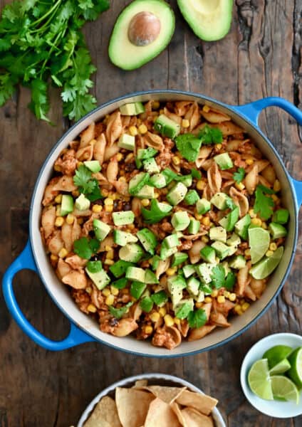 A dutch oven containing cheesy turkey taco pasta topped with diced avocado and chopped fresh cilantro next to a small plate with slices of limes.