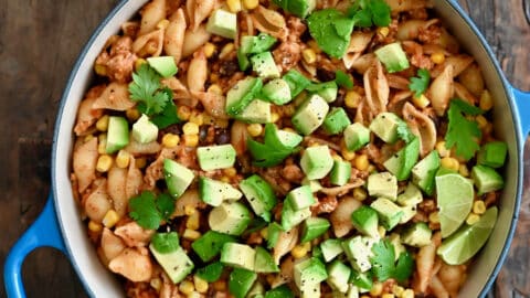 A dutch oven containing cheesy turkey taco pasta topped with diced avocado and chopped fresh cilantro next to a small plate with slices of limes.