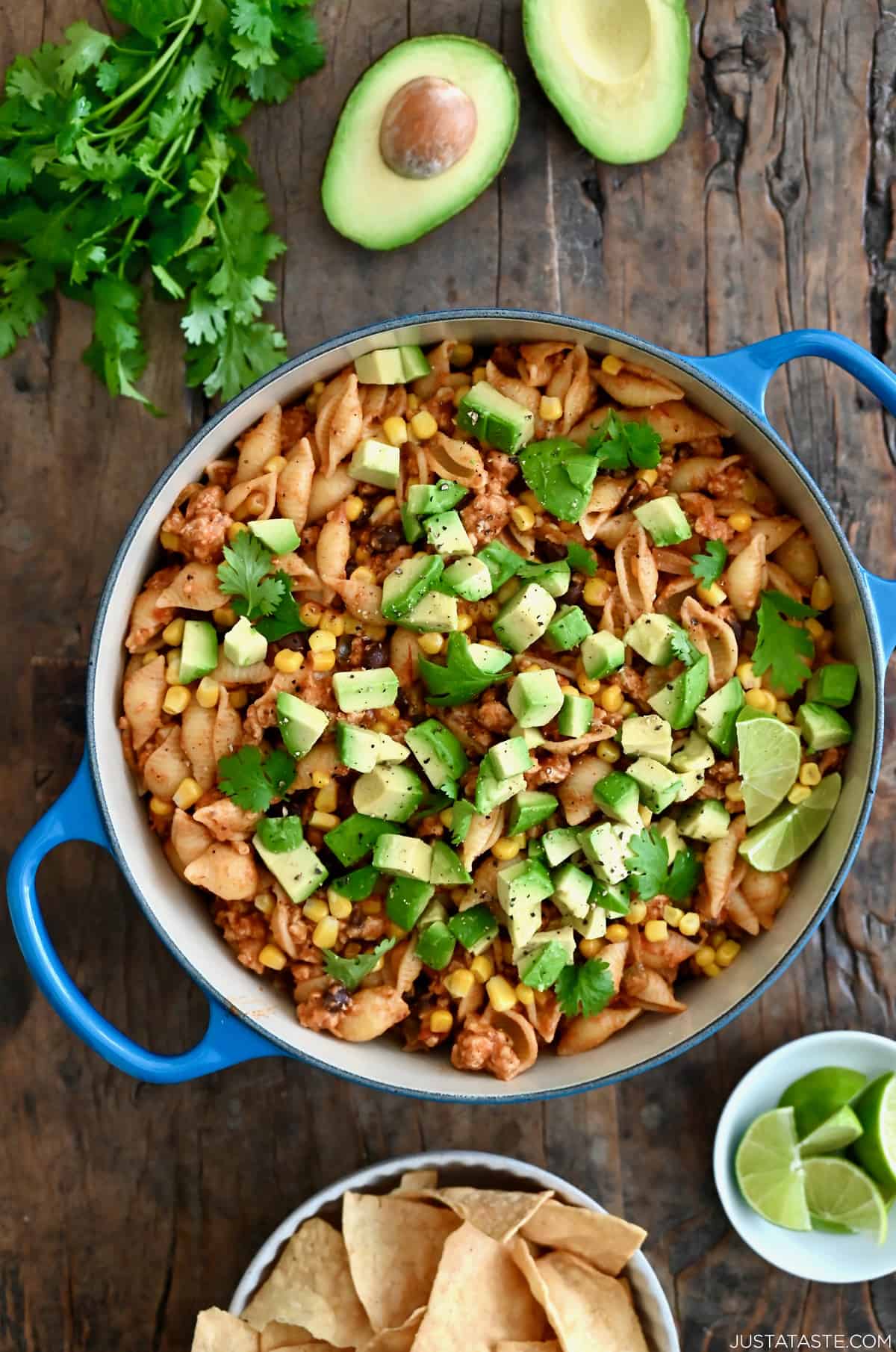 A dutch oven containing cheesy turkey taco pasta topped with diced avocado and chopped fresh cilantro next to a small plate with slices of limes.