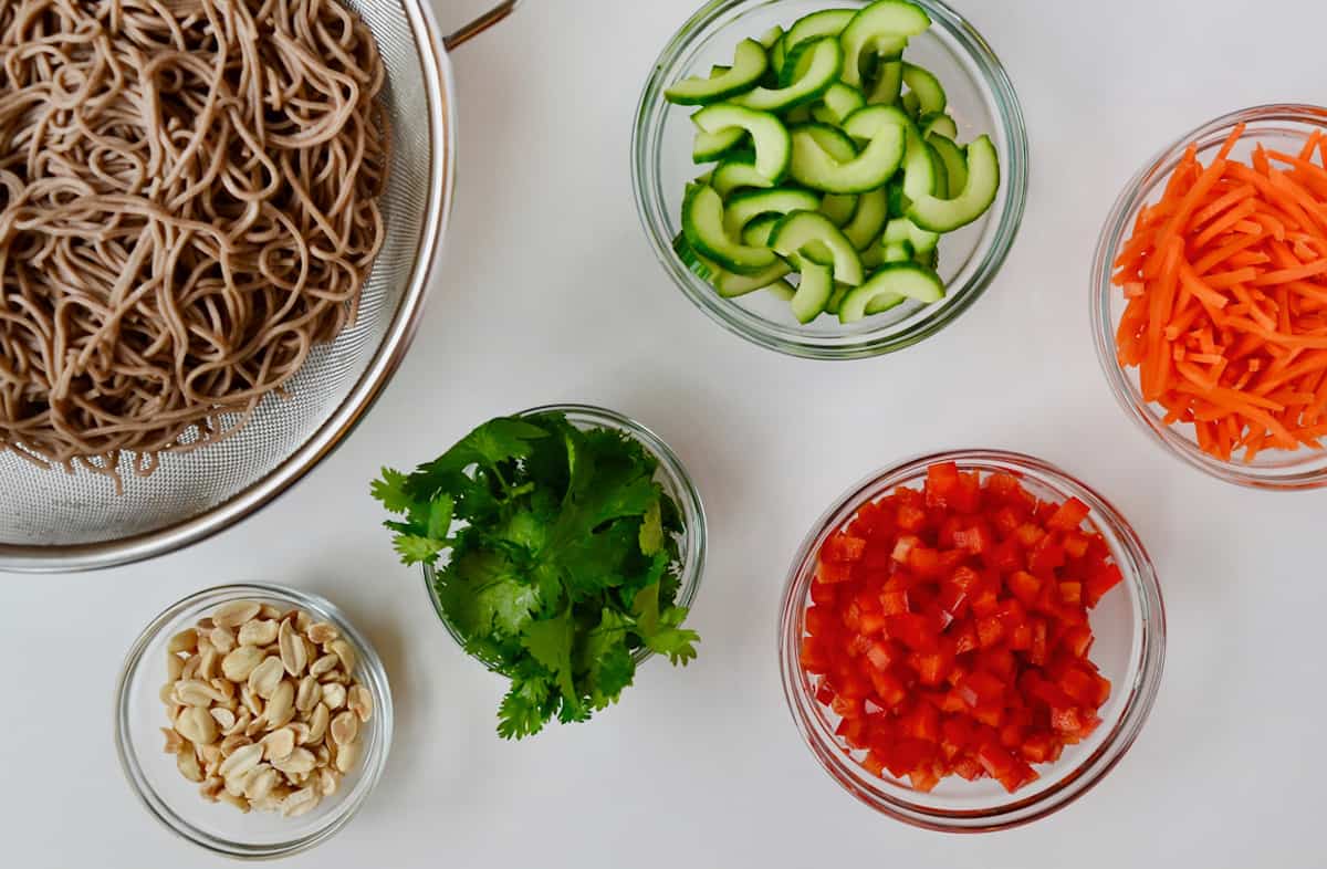 A colander full of cooked soba noodles and small bowls of peanuts, cilantro, diced red bell pepper, sliced cucumber and julienned carrots.