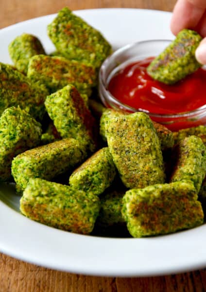 A platter of broccoli tots and a person dipping a baked broccoli tot into a small bowl of ketchup.