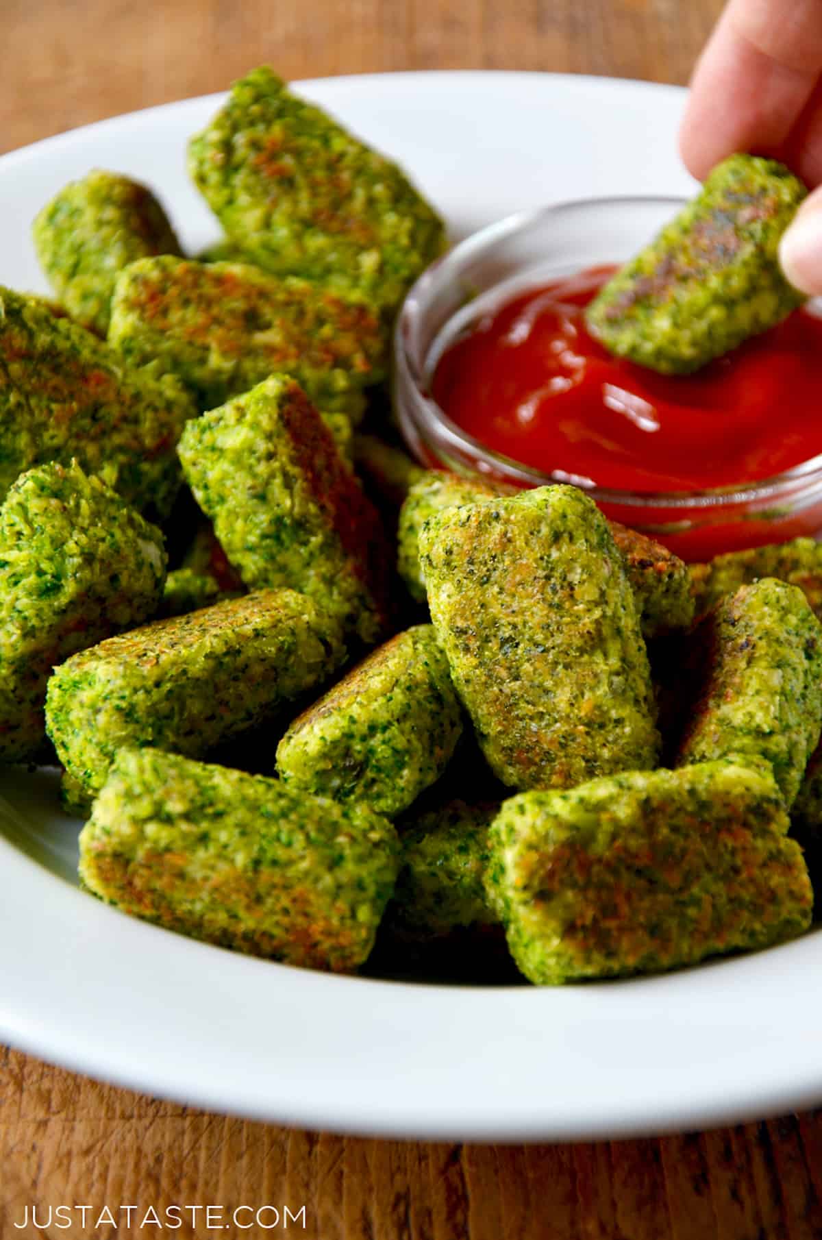 A platter of broccoli tots and a person dipping a baked broccoli tot into a small bowl of ketchup.
