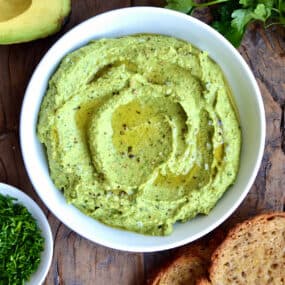 Avocado hummus in a bowl topped with olive oil, flakey salt and pepper. Small bowls of olive oil and minced chives, toasted bread, fresh parsley and a halved avocado surround the hummus.