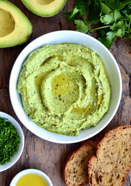 Avocado hummus in a bowl topped with olive oil, flakey salt and pepper. Small bowls of olive oil and minced chives, toasted bread, fresh parsley and a halved avocado surround the hummus.