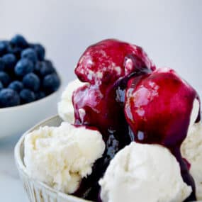 Blueberry sauce covering a bowl of vanilla ice cream. A small bowl with fresh blueberries sits behind the ice cream.