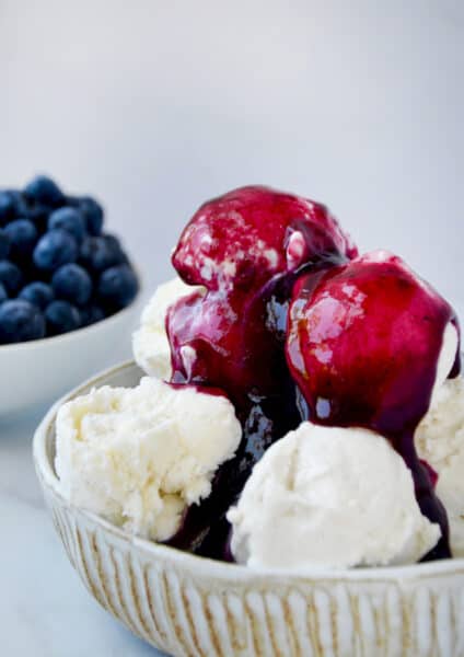 Blueberry sauce covering a bowl of vanilla ice cream. A small bowl with fresh blueberries sits behind the ice cream.