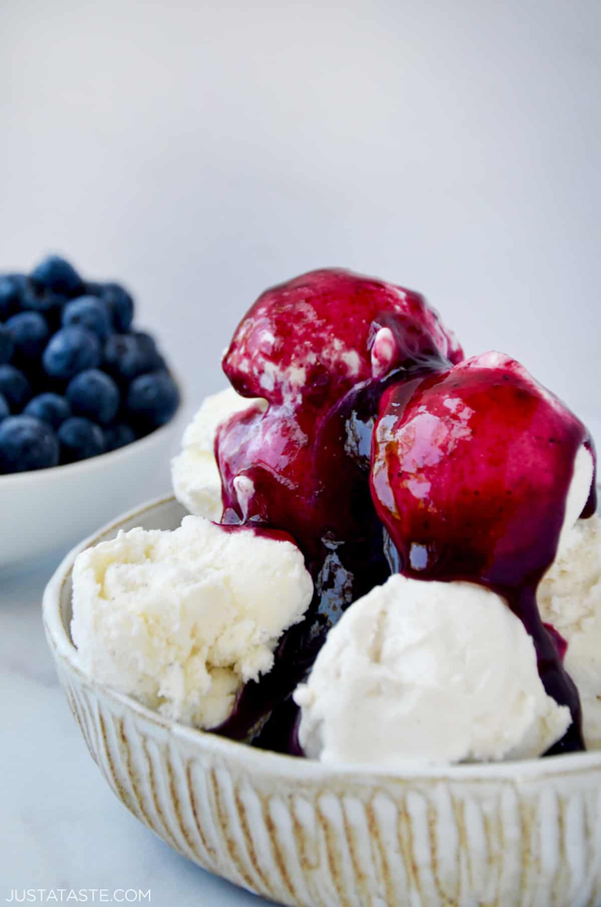 Blueberry sauce covering a bowl of vanilla ice cream. A small bowl with fresh blueberries sits behind the ice cream.