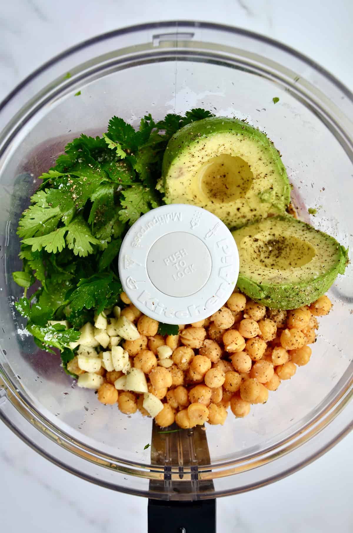 Chickpeas, chopped garlic, avocado halves, fresh herbs, salt and pepper in the bowl of a food processor.