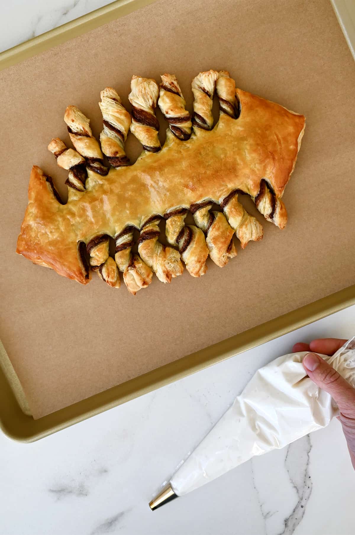 A football-shaped puff pastry with Nutella filling on a parchment paper-lined baking sheet next to a piping bag with vanilla frosting.